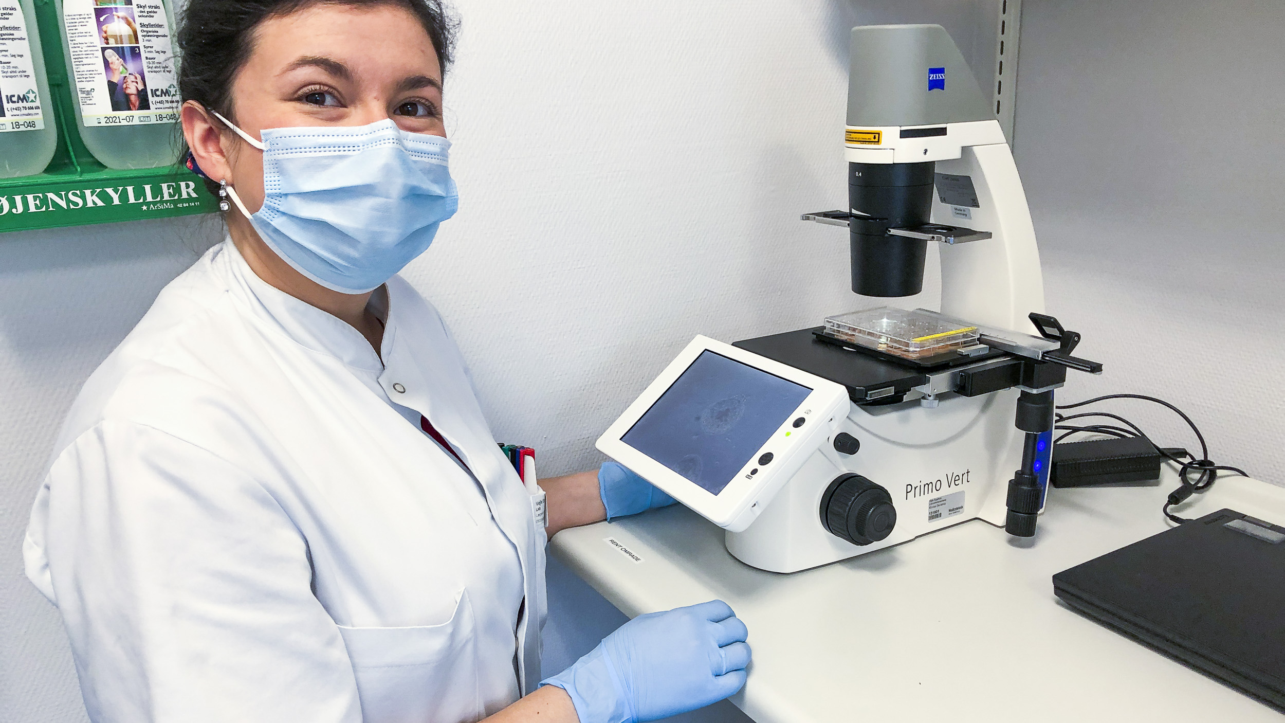 Sygeplejerske står foran mikroskop, hvor der ligger en prøve.